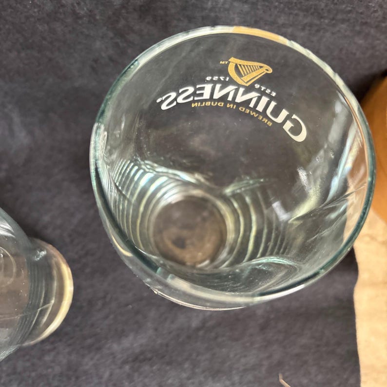 May include: Clear glass pint glass with the Guinness logo and text. The logo features a golden harp. The text reads "Guinness" and "Brewed in Dublin". The glass has a curved shape.