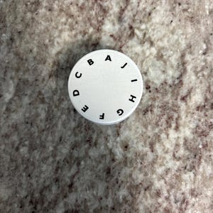 May include: A white, circular object with black letters A through J arranged around the edge. The object is set against a speckled, light brown and gray surface.