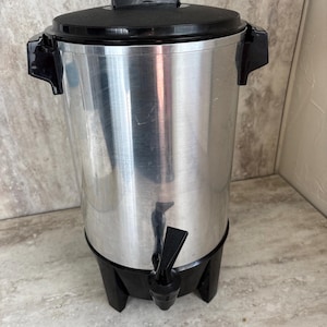 Puede incluir: Una cafetera de acero inoxidable con tapa, asas y base negras. La cafetera tiene un grifo para dispensar caf&eacute;. El dise&ntilde;o es sencillo y funcional, adecuado para servir caf&eacute; en eventos o en un entorno comercial.