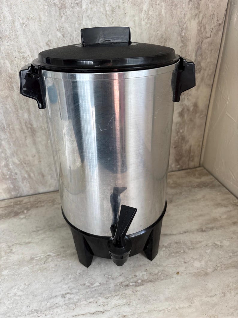 Puede incluir: Una cafetera de acero inoxidable con tapa, asas y base negras. La cafetera tiene un grifo para dispensar caf&eacute;. El dise&ntilde;o es sencillo y funcional, adecuado para servir caf&eacute; en eventos o en un entorno comercial.