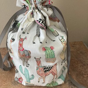 May include: A white drawstring bag with a gray ribbon. The bag is decorated with a pattern of llamas and cacti.