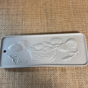 May include: A rectangular, light gray ceramic tray with a basket of fruit and wheat stalks embossed on the surface. The tray has a small hole on one end. The design is in a relief style.