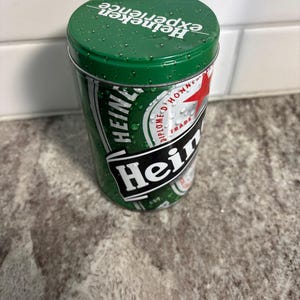 May include: A green Heineken tin with the words "Heineken experience" printed on the lid. The tin has a green and white design with the Heineken logo and other text. The cylindrical tin is made of metal.