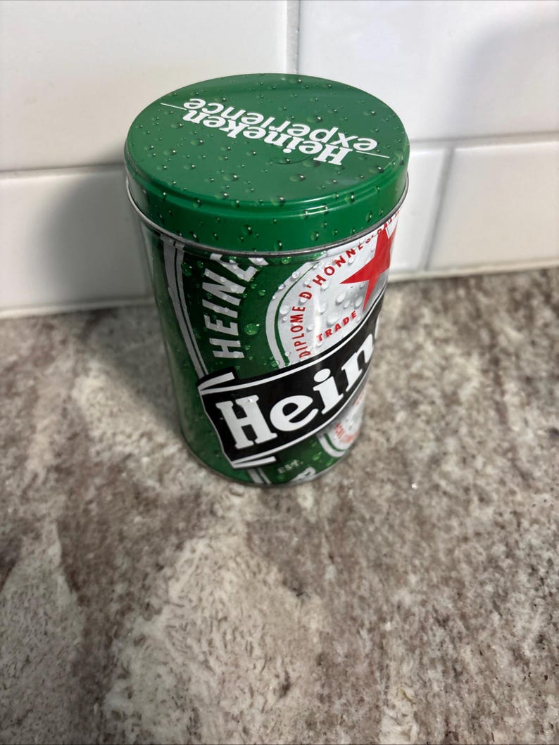 May include: A green Heineken tin with the words "Heineken experience" printed on the lid. The tin has a green and white design with the Heineken logo and other text. The cylindrical tin is made of metal.