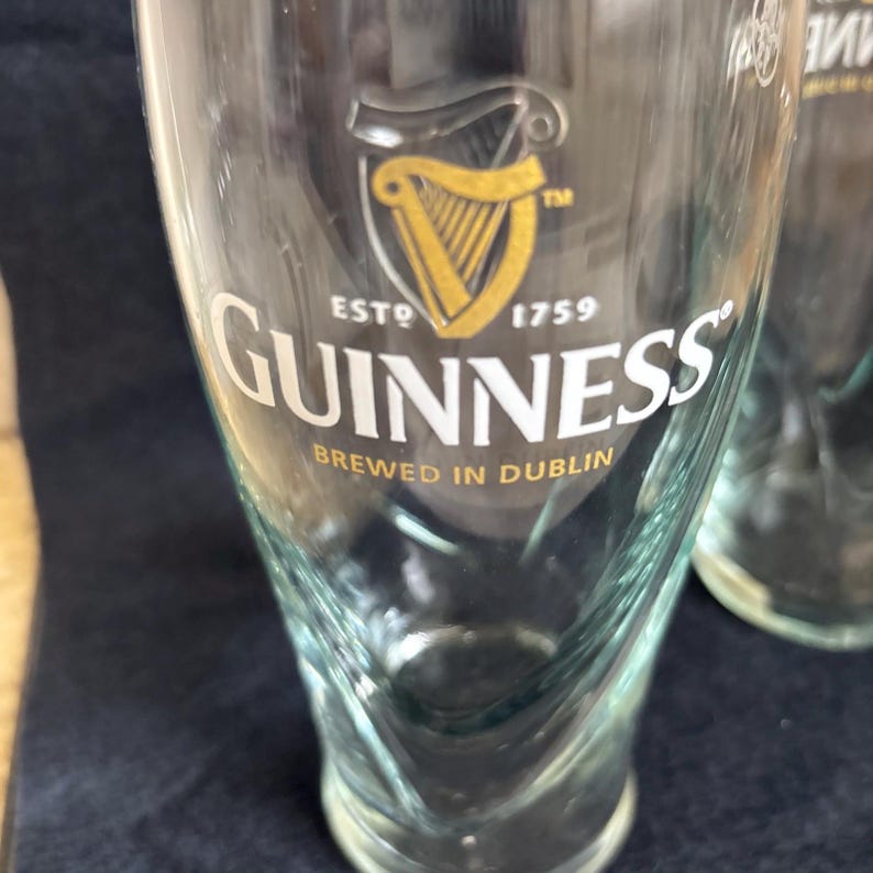 May include: Clear pint glass with the Guinness logo. The logo features a gold harp inside a black shield, with the word "Guinness" in white. The text "Brewed in Dublin" is below the logo.