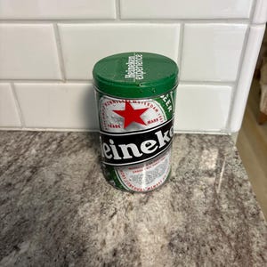 May include: A green and silver Heineken beer tin with a red star logo. The tin has the words "Heineken experience" on the lid and "Heineken" in large black letters on the side. The cylindrical tin sits on a speckled countertop.