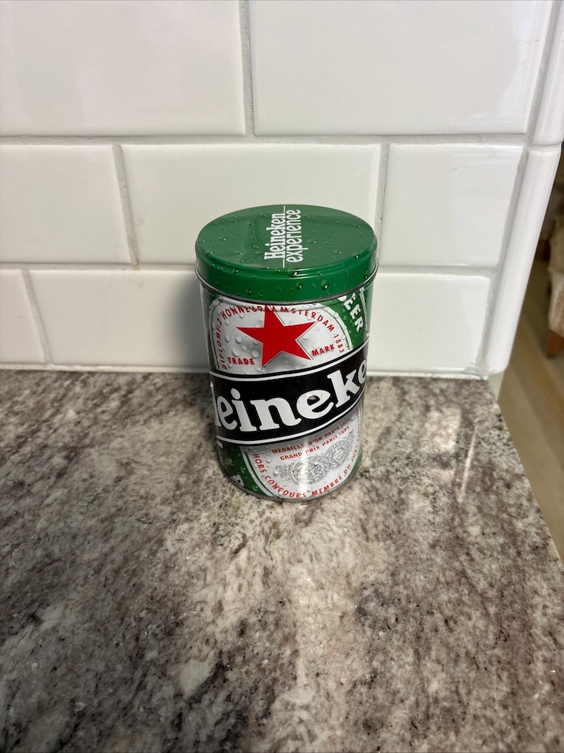 May include: A green and silver Heineken beer tin with a red star logo. The tin has the words "Heineken experience" on the lid and "Heineken" in large black letters on the side. The cylindrical tin sits on a speckled countertop.