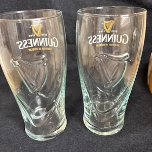 May include: Two clear Guinness pint glasses. Each glass features the Guinness logo with a harp and the text "Guinness" in white. The glasses have a curved shape and are designed for enjoying stout.
