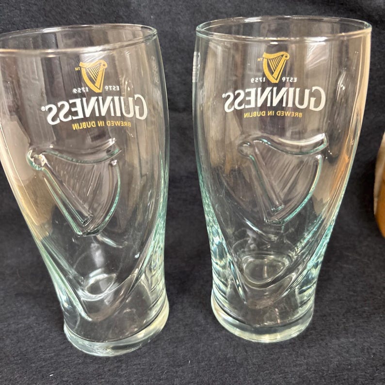 May include: Two clear Guinness pint glasses. Each glass features the Guinness logo with a harp and the text "Guinness" in white. The glasses have a curved shape and are designed for enjoying stout.