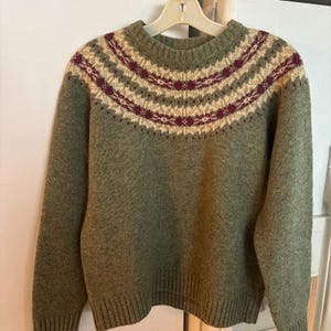 May include: A green sweater with a patterned yoke. The yoke features a repeating design of cream, burgundy, and green geometric shapes. The sweater has long sleeves and a ribbed hem and cuffs. The sweater is hanging on a white hanger.