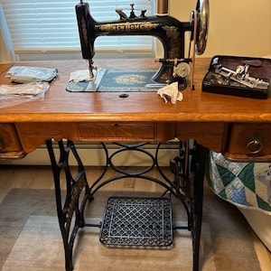 May include: An antique New Home sewing machine with a black and gold finish, set on a wooden table. The machine features intricate detailing and a large hand wheel. A foot pedal and a drawer are visible. The machine is a vintage item.