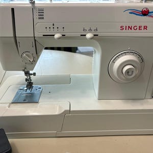 May include: A white Singer sewing machine featuring a horizontal control panel with stitch selection icons, two sliding adjustment levers, a circular tension dial, and a needle assembly. The brand name SINGER is printed in red on the front.