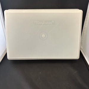 May include: A rectangular, off-white Tupperware container. The lid has the Tupperware logo and a circular indentation in the center. The container is made of a translucent plastic material.