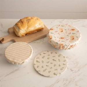 May include: Three reusable food covers made of natural fabric with different patterns. One cover has a striped pattern, another has a leaf pattern, and the third has a pattern of bread loaves and berries. The covers are on a white countertop next to a loaf of bread on a wooden cutting board.