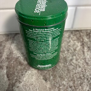 May include: A green Heineken souvenir glass container with the text "Heineken experience" on the lid. The container has text on the side describing the Heineken brewing process and the "Original Heineken Glass".
