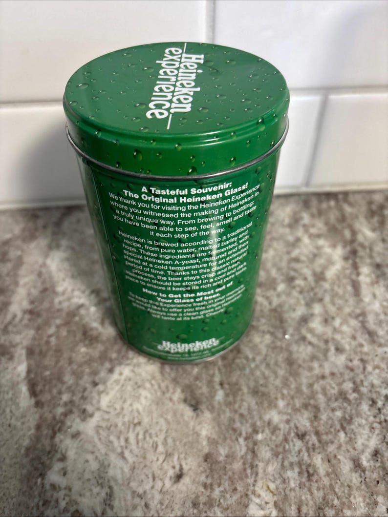 May include: A green Heineken souvenir glass container with the text "Heineken experience" on the lid. The container has text on the side describing the Heineken brewing process and the "Original Heineken Glass".