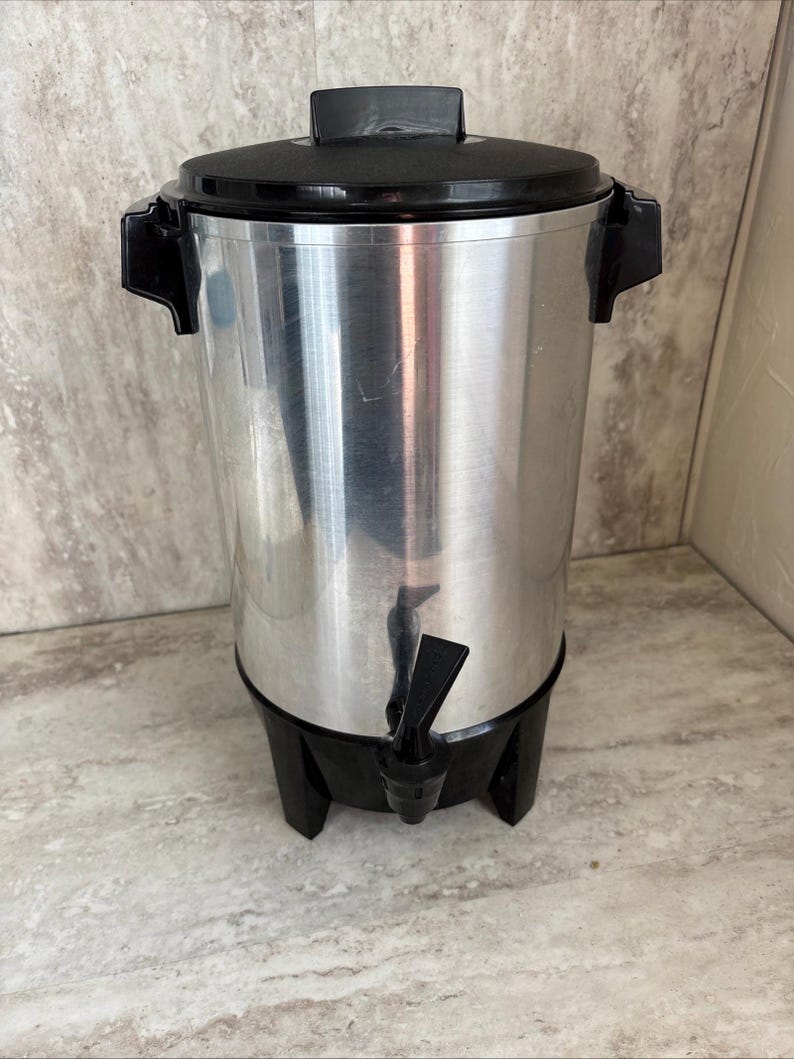 Puede incluir: Una cafetera de acero inoxidable con tapa, asas y base negras. La cafetera tiene un grifo para dispensar. El exterior est&aacute; pulido y es reflectante. Ideal para servir caf&eacute;.