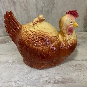 May include: Ceramic hen-shaped container with a chick on top. The container is primarily brown with yellow and red accents. The hen has a detailed feather design and a red comb. A decorative kitchen item.