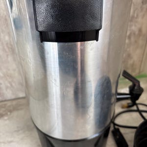 Puede incluir: Una cafetera antigua, cil&iacute;ndrica, de aluminio cepillado, con un asa y una base de pl&aacute;stico negro. La cafetera tiene un pico, un cable de alimentaci&oacute;n y una tapa negra. El aparato est&aacute; dise&ntilde;ado para preparar y servir caf&eacute;.