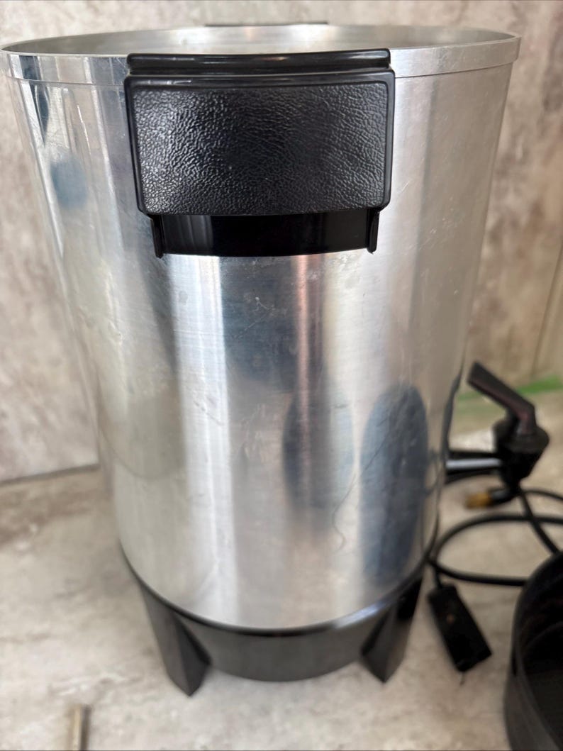 Puede incluir: Una cafetera antigua, cil&iacute;ndrica, de aluminio cepillado, con un asa y una base de pl&aacute;stico negro. La cafetera tiene un pico, un cable de alimentaci&oacute;n y una tapa negra. El aparato est&aacute; dise&ntilde;ado para preparar y servir caf&eacute;.