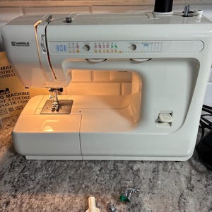 May include: A white Kenmore sewing machine with a black thread spool on top, featuring a stitch selection panel with letters A through I and length settings. Included are several metal presser feet, a screwdriver tool, and a small cleaning brush.