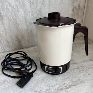 May include: A vintage electric coffee pot with a white body, brown lid, and handle. The base and trim are dark brown. A black power cord with a plug is coiled next to the pot. The text "Hot & Serve" is visible on the front.