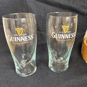 May include: Two clear Guinness pint glasses. Each glass features the Guinness logo with a harp and the text "ESTD 1759 Guinness BREWED IN DUBLIN". The glasses have a curved shape and are empty.