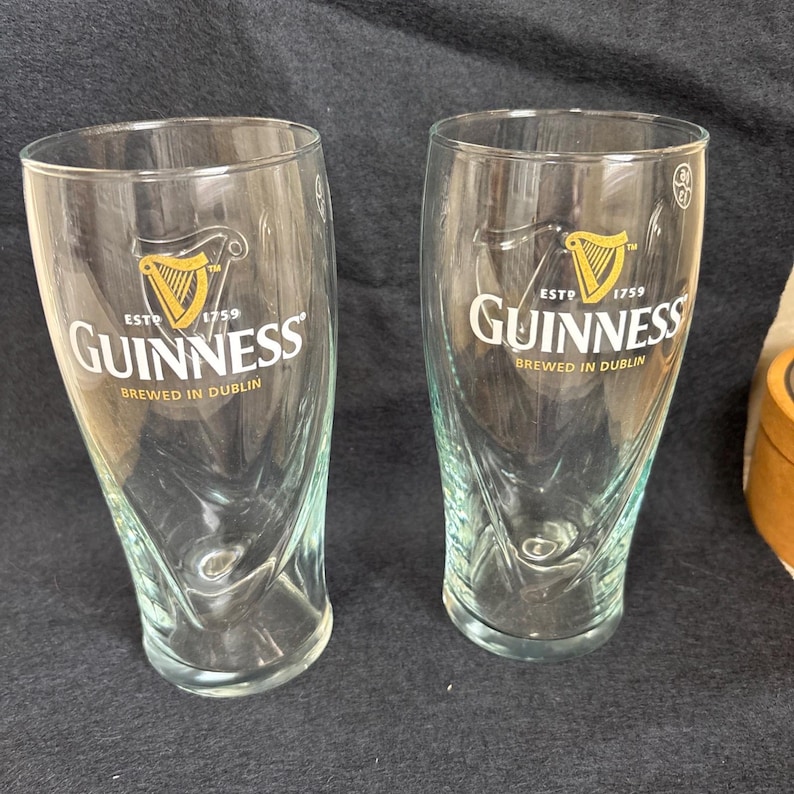 May include: Two clear Guinness pint glasses. Each glass features the Guinness logo with a harp and the text "ESTD 1759 Guinness BREWED IN DUBLIN". The glasses have a curved shape and are empty.