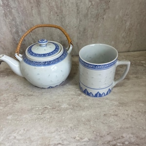 May include: White porcelain teapot and mug set with blue geometric patterns. The teapot has a woven handle and a lid with a floral design. The mug has a handle and a matching blue pattern.