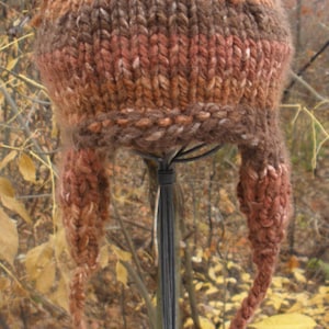 May include: A hand-knitted hat in shades of brown, rust, and cream, displayed on a wire stand. The hat features ear flaps and long braided ties with tassels. The textured knitwear is a cosy accessory.