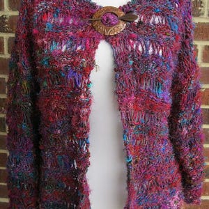 May include: A crocheted cardigan in shades of pink, purple, and blue with a wooden button closure.