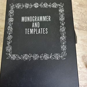 May include: A black binder with the words "MONOGRAMMER AND TEMPLATES" in white. The text is framed by a white floral border of roses and leaves. The binder is closed and appears to be on a light-colored surface.