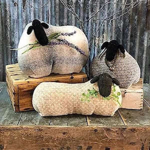 May include: Three handmade plush sheep in various poses. The sheep are made of a soft, textured fabric and have black faces and hooves. The sheep are sitting on a wooden crate.