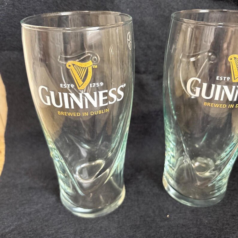 May include: Two clear Guinness pint glasses. Each glass features the Guinness logo with a gold harp and the text "ESTD 1759" and "BREWED IN DUBLIN". The glasses have a curved design and are made of clear glass.