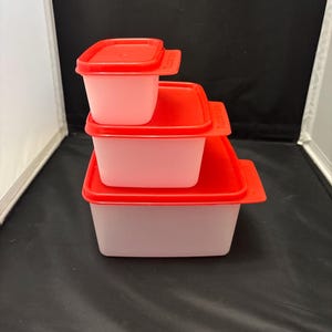 May include: A stack of three square food storage containers with red lids. The containers are different sizes, with the smallest on top and the largest on the bottom. The containers are a light pink color.