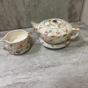 May include: A vintage floral tea set featuring a teapot, two cups, and a saucer. The set has a cream-colored base with a pattern of pink, yellow, and green flowers and foliage. The teapot has a lid and a handle, and the cups have handles.