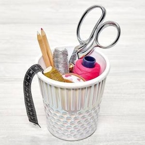 May include: A white thimble-shaped container holding sewing supplies including two wooden pencils, a spool of silver thread, a spool of hot pink thread, a spool of gold thread, and a pair of silver metal scissors with a black measuring tape.