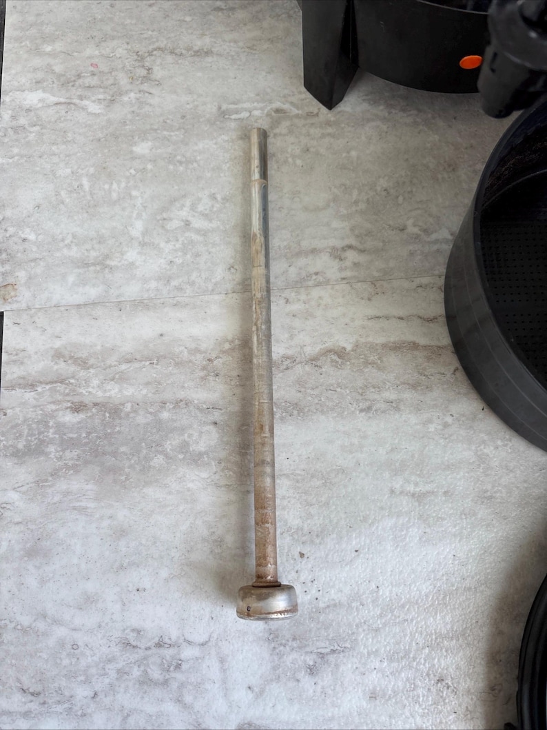 Puede incluir: Una larga varilla met&aacute;lica cil&iacute;ndrica con una base redondeada y ensanchada. La varilla tiene un aspecto ligeramente deslustrado y envejecido, lo que sugiere que puede ser vintage o usada. El objeto est&aacute; sobre una superficie clara y texturizada.