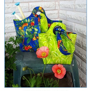 May include: Two reversible fabric bags with handles. The smaller bag is 6 inches by 6 inches by 17 inches and the larger bag is 9 inches by 9 inches by 21 inches. The bags are made of a blue and green fabric with a tropical print. The bags are sitting on a green table with a brick wall in the background. The text "Sisters' Common Thread Bindle Bag" is at the top of the image. The text "FULLY REVERSIBLE - TWO SIZES" is in the middle of the image. The text "Size: 6"X6"X17" and 9"X9"X21"" is below the text "FULLY REVERSIBLE - TWO SIZES". The text "Full-size pattern pieces included" is below the text "Size: 6"X6"X17" and 9"X9"X21"". The text "www.sisterscommonthread.com" is below the text "Full-size pattern pieces included". The text "Designed by Yvonne Cabella" is below the text "www.sisterscommonthread.com". The text "Item # SCT10163" is below the text "Designed by Yvonne Cabella".