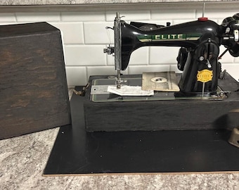 May include: A vintage black Elite sewing machine with green accents and a foot pedal. The machine sits on a black base, with a wooden carrying case to the left. A yellow sticker is on the machine.