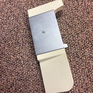 May include: A rectangular off-white plastic component featuring a central brushed metal plate. The plastic frame has a stepped design on one side and a rounded corner on the other. A small, circular, gear-like metal fastener is centered on the plate.