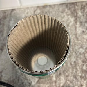 May include: A cylindrical container with a corrugated cardboard interior and a metal rim. The container is open at the top, revealing the textured interior. The exterior has a green and silver design.