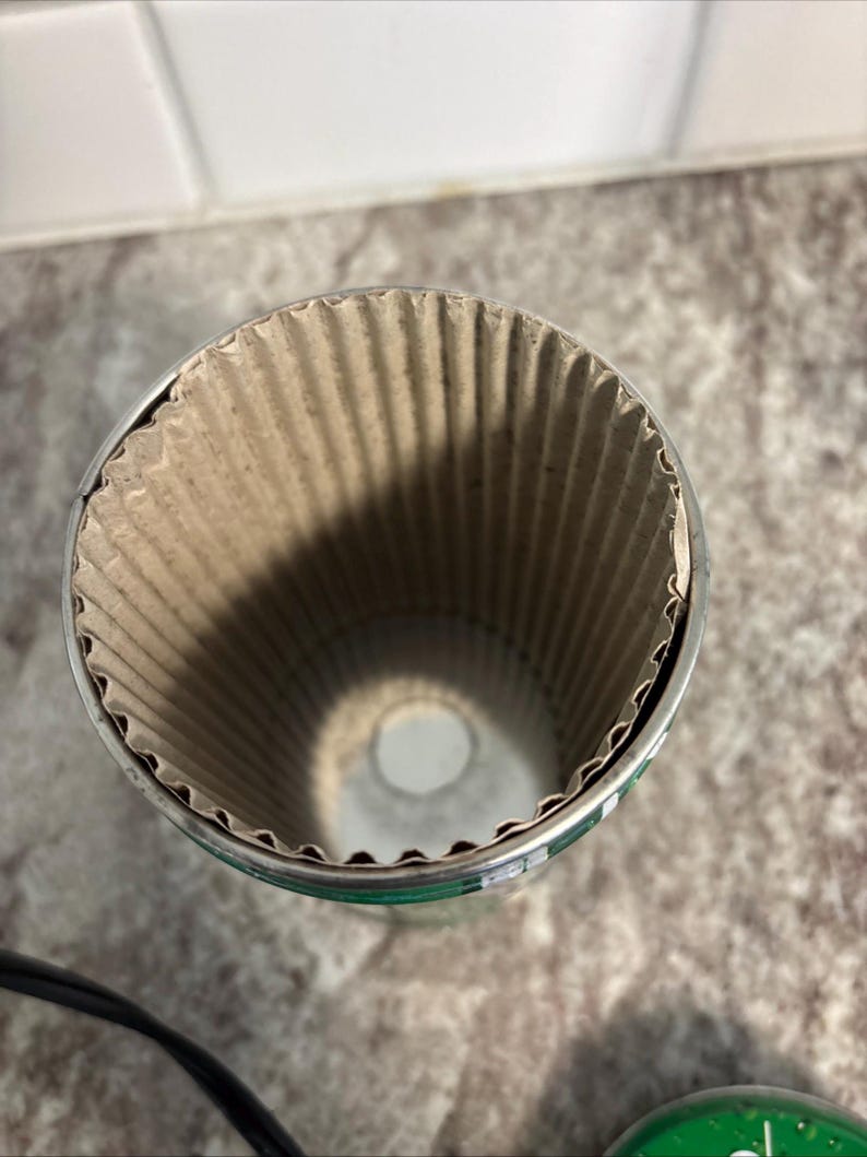 May include: A cylindrical container with a corrugated cardboard interior and a metal rim. The container is open at the top, revealing the textured interior. The exterior has a green and silver design.