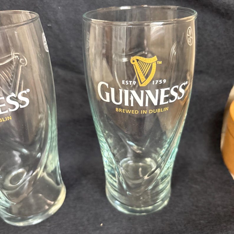 May include: Two clear pint glasses with the Guinness logo. The logo features a golden harp and the word "Guinness" in a bold, white font. The text "ESTD 1759" and "BREWED IN DUBLIN" are also visible on the glass.