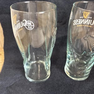 May include: Two clear Guinness pint glasses. Each glass has the Guinness logo and text. One glass has the numbers "06 13" printed on it. The glasses are on a dark blue surface.