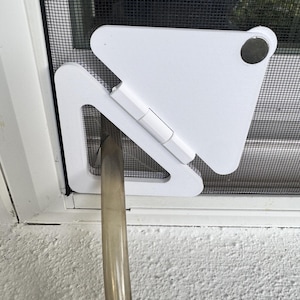 May include: A white plastic triangular pet door insert installed into a black mesh screen door. The device features a hinged flap mechanism and a circular silver-colored magnetic closure at the top corner, with a clear plastic hose passing through.