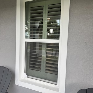 May include: A rectangular window with a white frame and white trim, featuring interior sage green plantation shutters with horizontal slats. The window is set into a gray exterior wall, with a small white corner piece visible at the bottom left.