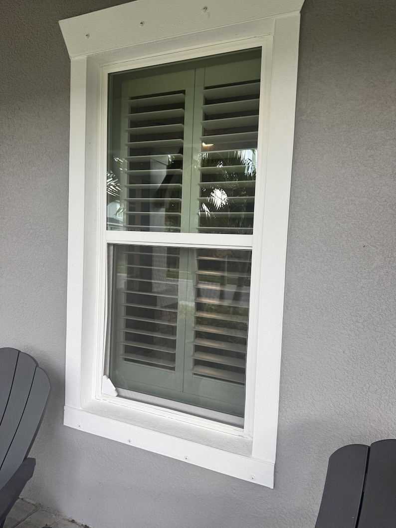 May include: A rectangular window with a white frame and white trim, featuring interior sage green plantation shutters with horizontal slats. The window is set into a gray exterior wall, with a small white corner piece visible at the bottom left.