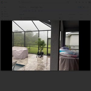 May include: A covered patio with a view of a rainy day. The patio has a table and chairs covered with a light-colored tarp. A stationary bike and a small tree are also visible. In the background, there is a fence and a house.