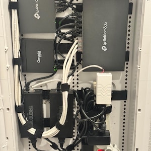 May include: A network equipment setup featuring multiple black TP-Link Omada and TRENDnet devices, along with various cables and power adapters, all neatly organized within a white enclosure. The setup includes a power strip and a small circuit board.